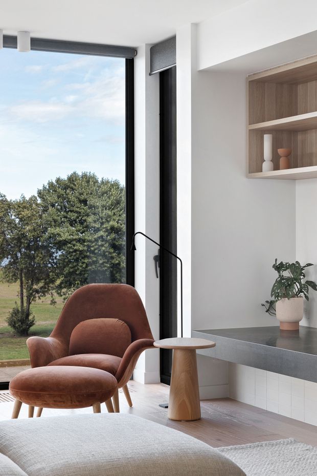 A bright, contemporary interior corner combining a living area with a compact kitchen or built-in workspace. Floor-to-ceiling black-framed glazing overlooks trees and lawn. A sculptural rust-brown lounge chair and ottoman sit beside a small timber side table and slim black floor lamp. To the right, a dark grey benchtop with white tiled cabinetry is paired with an open timber display shelf, plant, and ceramic objects. Pale timber flooring, white walls, and soft neutral furnishings create a refined, warm space.