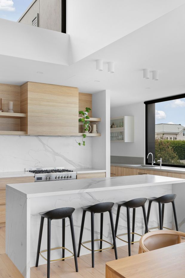 Bright contemporary kitchen with a large white marble-look waterfall island and seating for five, pale timber cabinetry, open shelving, white walls and ceiling, black-framed glazing, and dark bar stools with brass footrests. A compact dining table and chair are visible in the foreground, while the sink and additional cabinetry sit by the window.