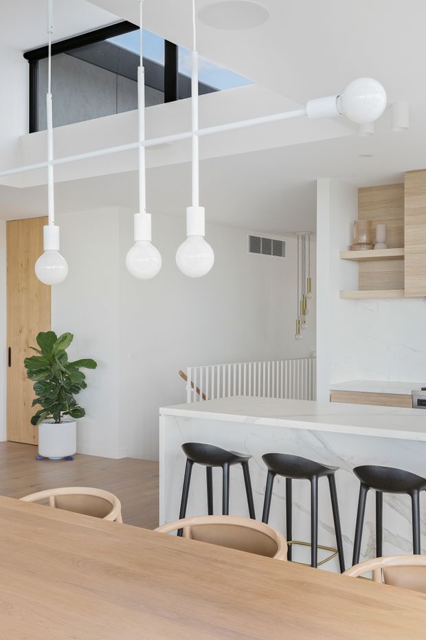 Bright open-plan kitchen and dining area with a white marble-look island, black bar stools, pale timber cabinetry and shelving, a light timber dining table, and sculptural white pendant lights. A stair balustrade and partial upper-level void suggest a contemporary multi-level interior.