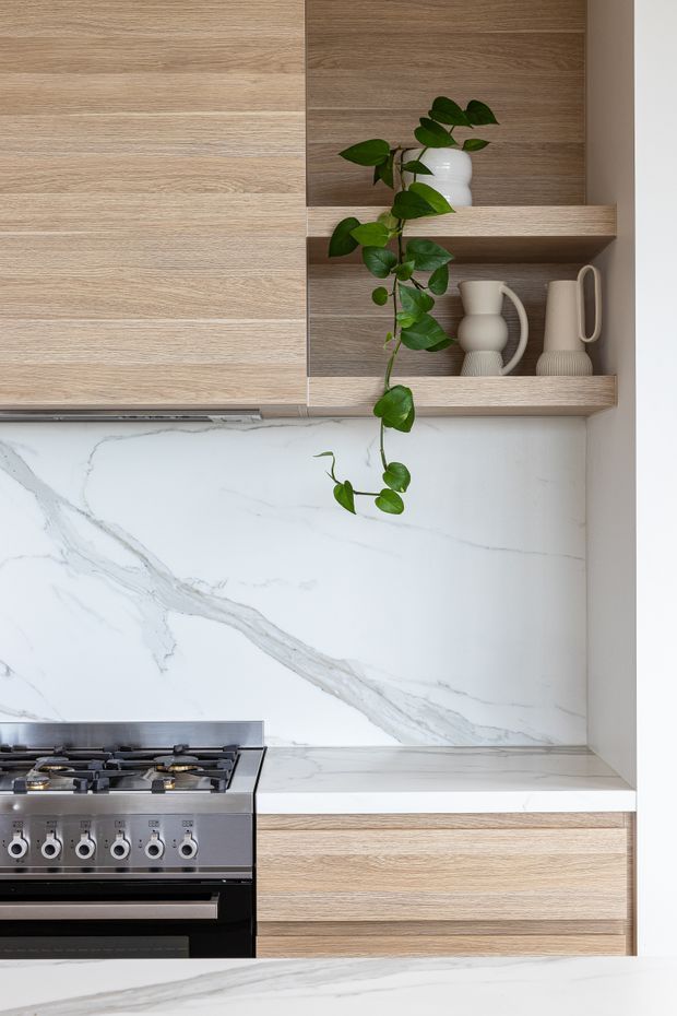 Close-up view of a refined contemporary kitchen featuring light timber cabinetry, open timber display shelving, a dramatic white marble-look splashback and benchtop with grey veining, and a stainless steel and black freestanding gas range. Neutral ceramic vessels and a trailing green plant add soft decorative accents.