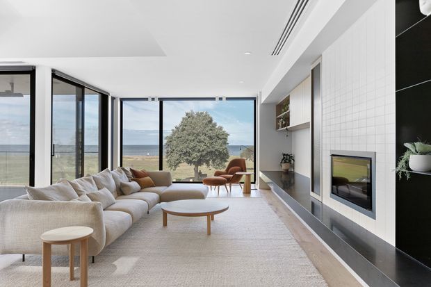 A bright, spacious contemporary living room with a large cream sectional sofa, pale textured rug, and low timber coffee and side tables. Full-height glazing opens to a coastal ocean view, while a built-in white tiled fireplace wall with dark stone hearth and integrated shelving forms the main feature. A sculptural lounge chair and indoor plants add warmth to the calm neutral interior.