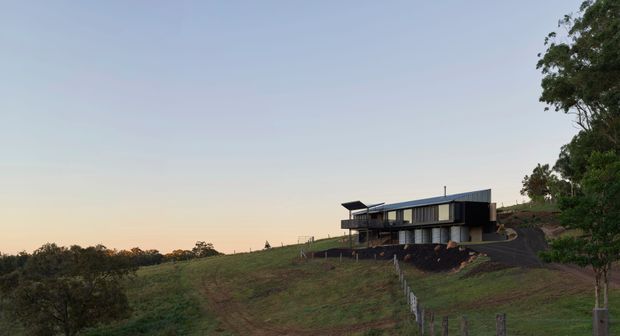 A contemporary dark-clad house sits on a gently sloping rural hillside, with broad horizontal glazing, projecting roof elements, and elevated structural supports. The property is surrounded by open grassland and scattered trees, viewed in soft early-evening light.