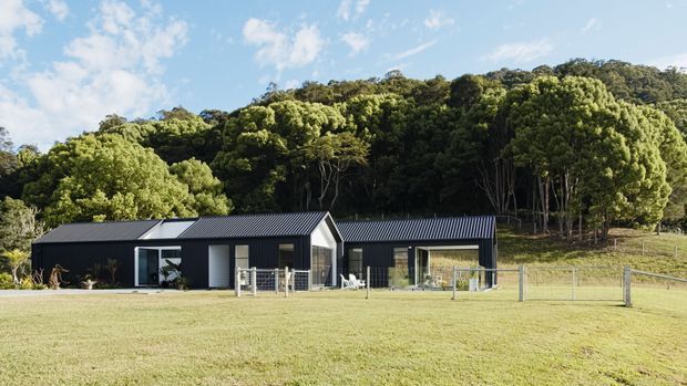 A low, single-storey contemporary rural residence with dark corrugated metal cladding and pitched gabled roofs, set in a broad lawn at the edge of dense native bush. Large glazed openings and a small fenced terrace face the open field, while the wooded hillside rises behind the house.