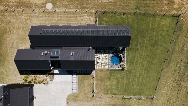 Aerial view of a contemporary dark-clad single-storey property on a large grassy site. The building has long rectangular wings with dark pitched or gently sloping roofs, visible rooftop solar panels and skylights, a concrete driveway, stepping-stone paths, and a small round blue swimming pool within a paved outdoor area. Wire fencing encloses the lawn.