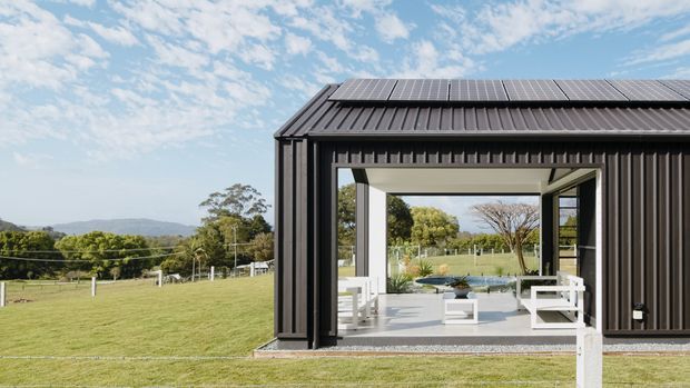 Exterior view of a dark, modern single-storey pavilion or outbuilding set within a spacious rural landscape. The building has black vertical metal cladding, a pitched corrugated metal roof fitted with solar panels, and a large open glazed frontage leading to a bright outdoor living area with white seating. Fencing, lawn, trees and distant hills are visible around the structure.