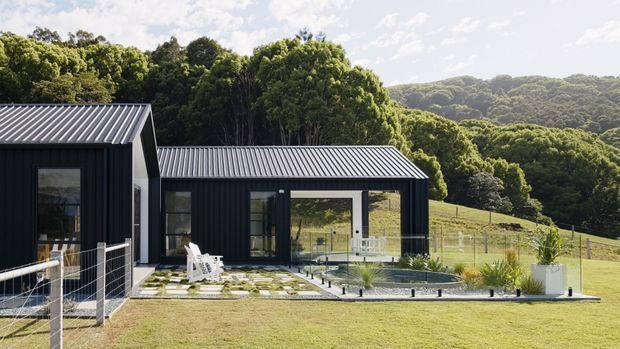 Exterior view of a modern single-storey house set in a lush, wooded sloping landscape. The building has dark vertical cladding, sharp gabled roof forms with corrugated metal roofing, large glazed openings, and a small paved terrace beside a narrow reflecting pool or water feature. Wire fencing and open lawn surround the home.