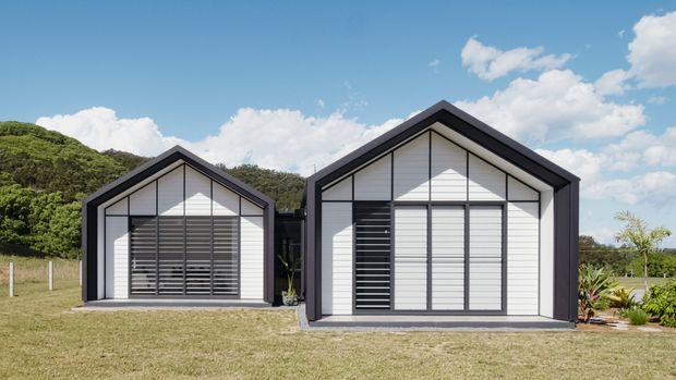Exterior view of two adjacent small gable-roofed buildings with crisp white horizontal cladding, dark charcoal framing and trim, and large glazed openings fitted with horizontal dark louvres. The buildings sit on a grassy landscaped site with wooded hills behind them under a bright blue sky.