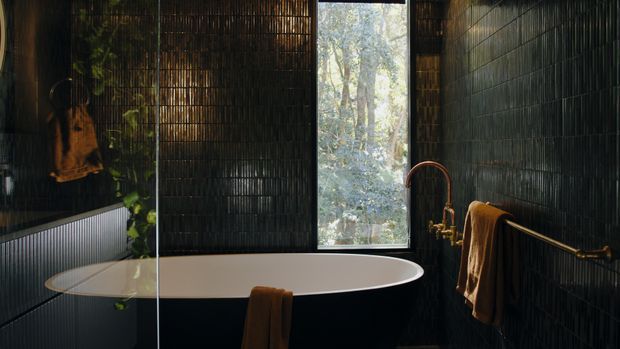 A moody, refined bathroom featuring a freestanding white oval bathtub against dark vertically textured wall tiles. A tall narrow window brings in filtered views of trees, while warm brass tapware and towel rails add contrast. Dark built-in cabinetry and a trailing plant are visible at the side.
