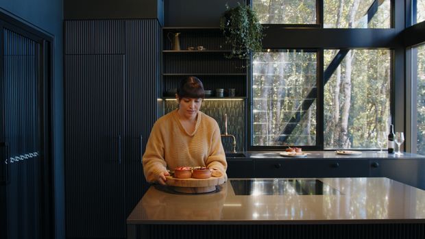 A dark, sophisticated contemporary kitchen with a large polished stone island in the foreground. A person stands at the island holding a wooden serving board, while black built-in cabinetry, vertically fluted wall panels, open shelving, integrated lighting, dark-framed glazing, and a forest view define the space.