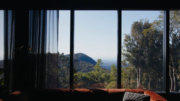 Interior view through large floor-to-ceiling windows, with dark window frames and sheer curtains framing a mountainous, tree-covered landscape. The lower foreground shows part of a deep rust-toned sofa and a textured cushion, creating a moody, intimate viewing space.