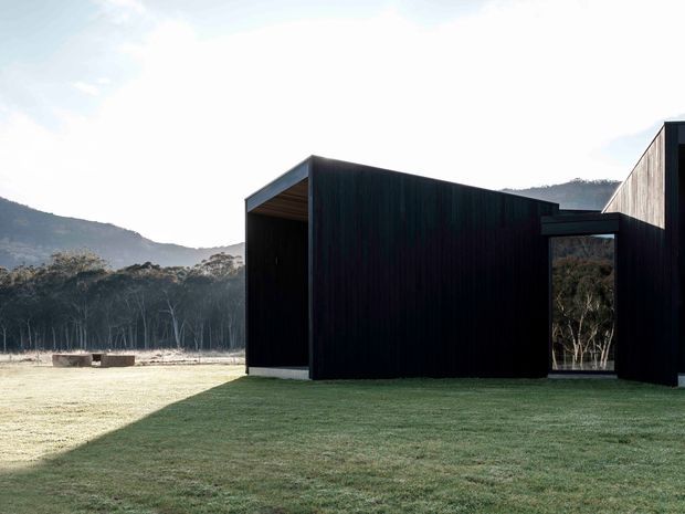 Exterior view of a minimalist, low-profile building set in a rural or bush landscape. The structure has dark vertical cladding, strong rectilinear volumes, a deep open recessed entry or patio, and large glazed openings. A broad lawn and surrounding wooded hills are visible.