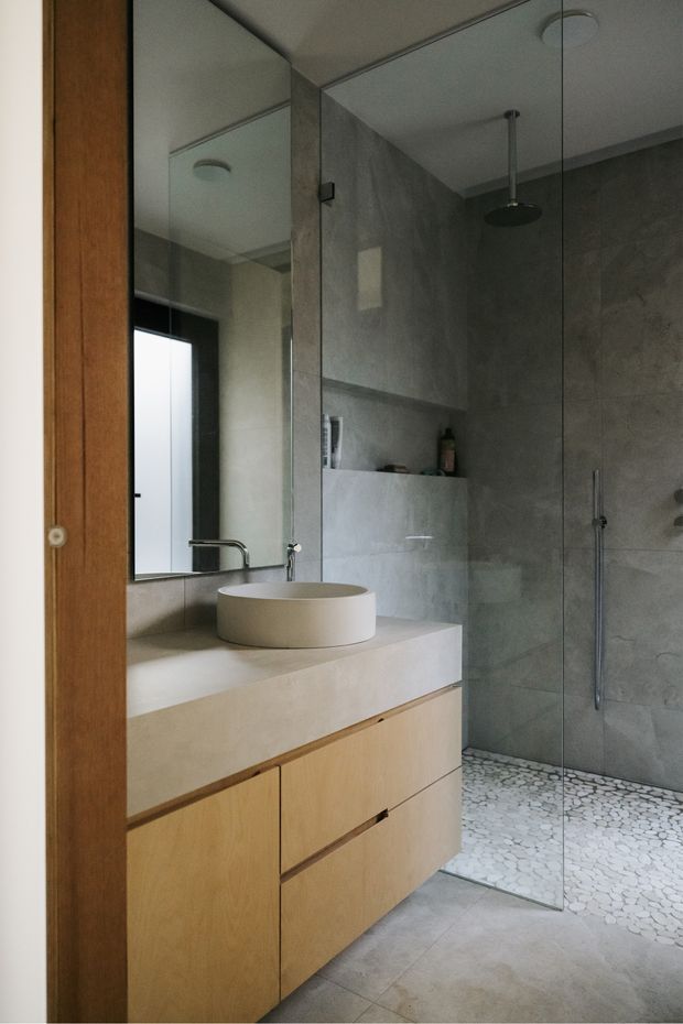 A compact contemporary bathroom with a floating light timber vanity, integrated pale stone or concrete benchtop, round vessel basin, wall-mounted tap, large mirror, and frameless glass walk-in shower. The shower walls have a grey textured concrete-look finish, with a recessed shelf and dark shower fittings. Small light pebble-style tiles define the shower floor, while large pale grey floor tiles continue through the vanity area. A timber-framed doorway is visible at left.