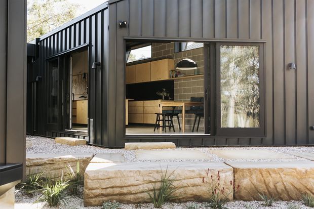 Exterior view of a dark, single-storey contemporary building clad in vertically ribbed metal panels. Large glazed openings reveal a warm kitchen and dining area inside, while the entrance and facade sit above layered natural-stone landscape steps with gravel and native planting.