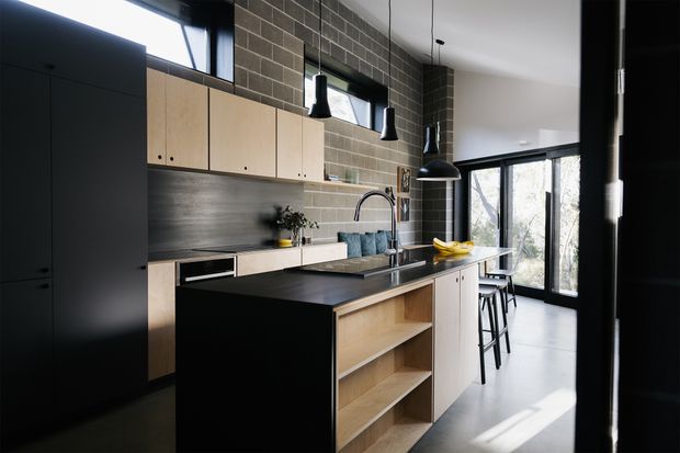 Contemporary kitchen with a large dark island, pale timber cabinetry, integrated dark appliances, a stainless-steel splashback, and a textured dark brick feature wall. Black-framed windows and pendant lights create strong contrast, while a breakfast bar with stools sits beside the glazing.