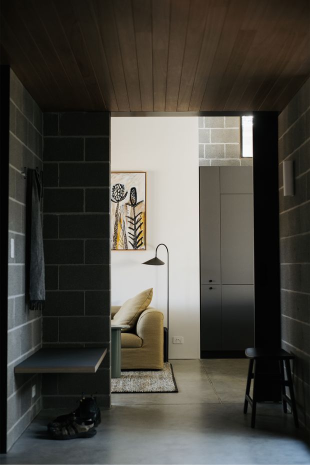 A dark, narrow entry hall framed by charcoal-painted concrete block walls and a warm timber-lined ceiling. The hall opens into a bright living room with a pale upholstered sofa, floor lamp, artwork, built-in dark cabinetry and a textured rug. A bench, stool and shoes reinforce the entryway function. The space has a moody, modern-industrial character with strong contrast between the dark corridor and light living area.