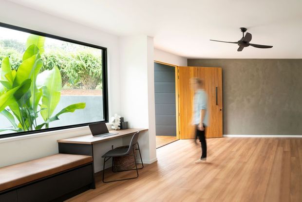 Bright contemporary open-plan interior featuring a timber-floored living or entry area, a large black-framed window with leafy planting outside, built-in timber bench seating, a compact desk, and a striking timber pivot entry door beside dark wall panelling. A ceiling fan and textured grey feature wall add visual interest.