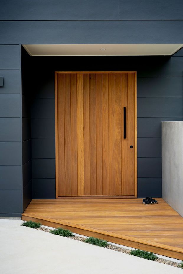 Modern residential entrance featuring a large vertical timber pivot door set within a recessed black entry alcove. The facade is clad in dark horizontal panels, with a warm timber deck and threshold, concealed soffit lighting, and a minimalist concrete wall at the side.