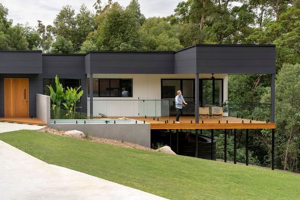A modern single-storey house set into a leafy, sloping site. The dark horizontal-clad facade has a flat roof, warm timber entry door and elevated timber terrace with clear glass balustrading. Large glazed openings connect the interior to the deck and surrounding bush landscape, with a partially visible lower level beneath the terrace.