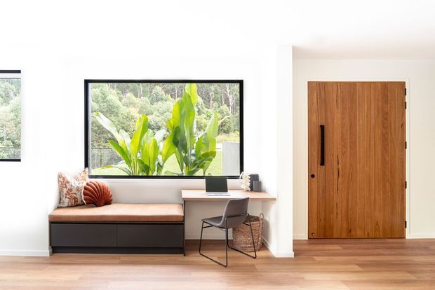 Bright contemporary entry area with a built-in window seat, compact home workstation, timber flooring, large black-framed window, and a tall natural timber entry door. Lush greenery is visible outside the window.