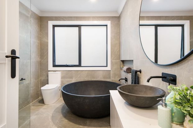 A refined contemporary bathroom featuring a freestanding dark stone bathtub beneath a large window, a matching vessel basin on a white vanity, warm grey stone-look wall and floor finishes, matte black tapware and window frames, and a large round mirror. A partially open door provides direct visual access into the bathroom.