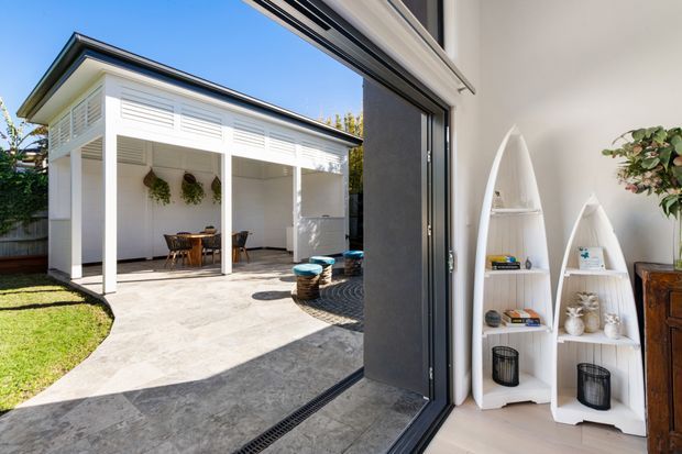 Bright contemporary indoor-outdoor living area opening onto a covered courtyard patio. The white pavilion has louvred upper wall panels, slim columns and a dark-edged low-pitched roof, with an outdoor dining setting beneath. Inside, white surfboard-shaped display shelves, potted greenery and a timber cabinet add coastal character. Large folding or stacking glass doors connect the interior to the paved outdoor area.