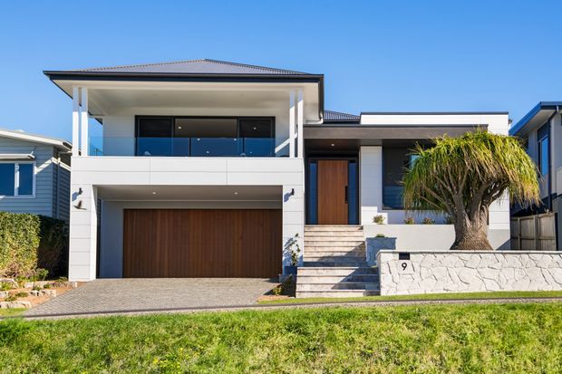 Contemporary two-storey suburban residence with a low-pitched hipped roof, white rendered facade, dark-framed glazing, timber garage door and entry door, and a raised landscaped front garden with broad steps and retaining wall.