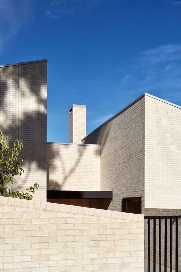 Exterior architectural photograph of a modern multi-level building composed of pale cream brick volumes with crisp parapets and pitched roof forms. A tall brick chimney, recessed dark entry canopy, timber door, foreground brick boundary wall, and black metal gate are visible. Strong sunlight creates tree shadows across the masonry against a clear blue sky.