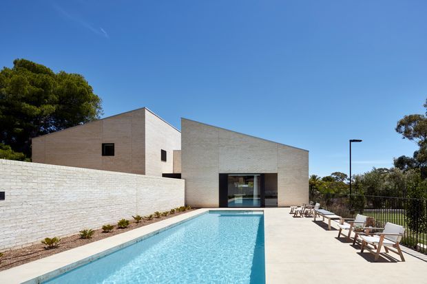 A contemporary low-rise residential building viewed from an outdoor pool terrace. The house has simple intersecting cubic volumes clad in pale masonry, a large dark-framed glazed opening, and a long rectangular swimming pool bordered by light paving. A white masonry boundary wall, sparse planting, outdoor lounge chairs, black metal balustrading, and mature trees frame the scene under a clear blue sky.