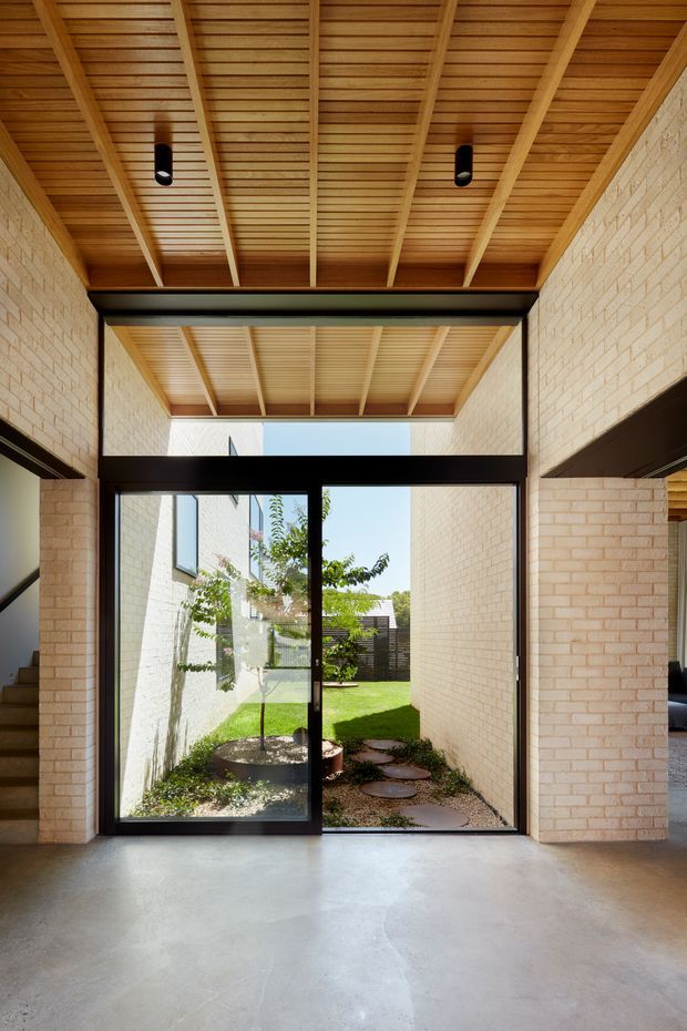 Interior view of a bright, high-ceilinged circulation or entry space opening onto a small landscaped courtyard. The space features exposed timber ceiling battens and beams, pale brick walls, black steel-framed glazing and structural beams, and a smooth polished concrete floor. A stair is partially visible at left and a glimpse of an adjoining living area appears at right.