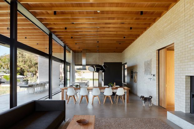 A spacious contemporary open-plan living and dining area with full-height black-framed glazing, a warm timber slatted ceiling with exposed beams, pale brick walls, and a long dining table with white chairs. A dark sofa, timber coffee table, fireplace, and indoor plants complete the refined, airy interior, with a landscaped outdoor area visible through the glazing.