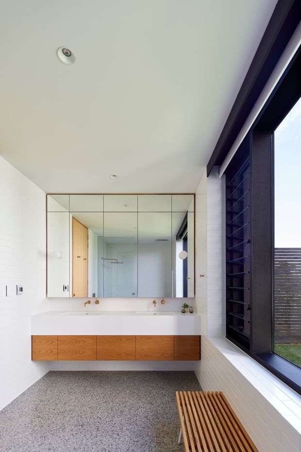 Bright contemporary bathroom with a double-sink floating timber vanity, white integrated benchtop, large mirrored wall cabinet, white tiled walls, terrazzo-style flooring, and a full-height dark-framed louvred window. A timber slatted bench sits below the window, and a shower area is visible in the mirror reflection.