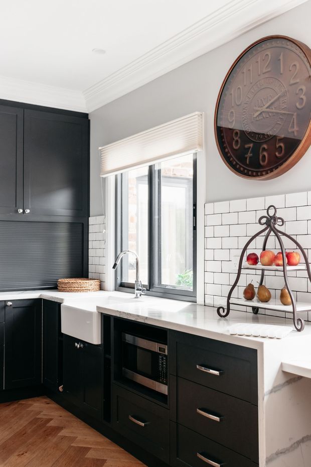 A bright, refined kitchen with matte black shaker-style cabinetry, a white farmhouse sink beneath a window, white subway-tile splashback, and a white stone benchtop. The room includes brushed metal hardware, an integrated microwave, a large vintage wall clock, and a decorative metal fruit stand. Warm herringbone timber flooring contrasts with the black-and-white cabinetry.