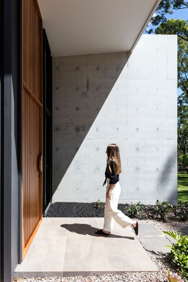 A contemporary residential entryway viewed from outside, featuring a tall timber slatted entry door, dark-framed glazing, a smooth exposed-concrete wall, and a broad white concrete canopy or wall plane. Strong sunlight creates a sharp diagonal shadow across the facade and pale stone-paved path, with a small landscaped garden at the edge.
