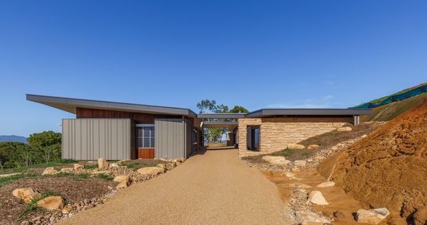Single-storey contemporary residential building viewed from a gravel driveway, integrated into a sloping landscaped site. The structure has low-pitched projecting roof planes, dark vertical metal cladding, warm timber wall sections, extensive glazing, and a substantial tan stone-clad wing partially embedded into the hillside. Clear blue sky and surrounding vegetation are visible in the background.