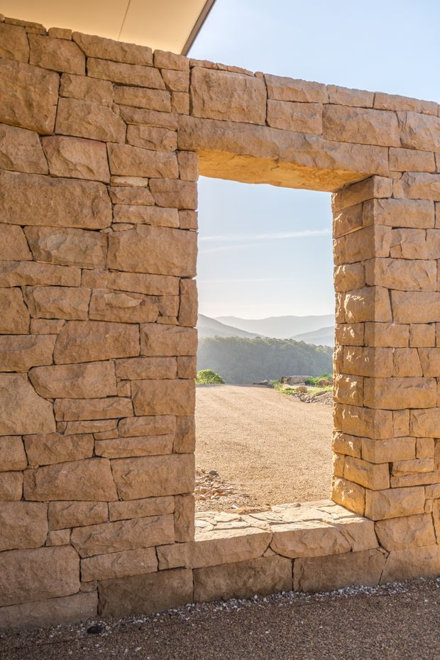 A close-up architectural view of a thick, rough-stacked natural stone wall with a rectangular, unglazed opening. The opening frames a sunlit gravel landscape and distant mountain ridges, while a pale overhanging roof or canopy is visible at the top. The scene has a warm, rugged, rustic character with strong natural textures and earthy tones.