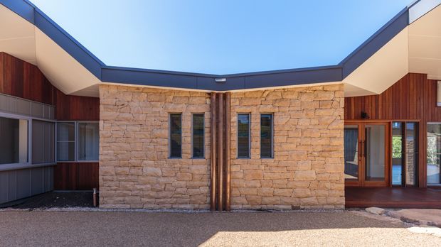Front-facing exterior view of a low, single-storey building arranged around a central stone-clad entry volume. The facade combines warm rough-cut masonry, dark vertical timber cladding, large glazed openings with timber frames, and a sharply angled dark roofline beneath a clear blue sky.