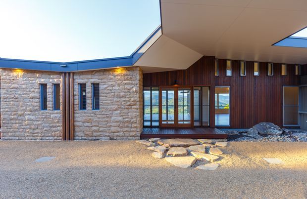 Exterior view of a contemporary single-storey building arranged around a sheltered entry or courtyard. The facade combines pale rough stone masonry, dark vertical timber cladding, narrow timber-framed windows, broad glazed doors, and a projecting flat roof with a blue-edged fascia. Gravel paving, stone stepping stones, and a skylight-like roof opening are visible.