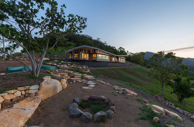 A modern single-storey home set on a steep, landscaped hillside at dusk. The building has extensive warm-lit glazing, dark vertical cladding, a low pitched roof, and a long linear form following the slope. Natural stone retaining edges, stepped paths, planted terraces, mature trees, and distant wooded hills surround the house.