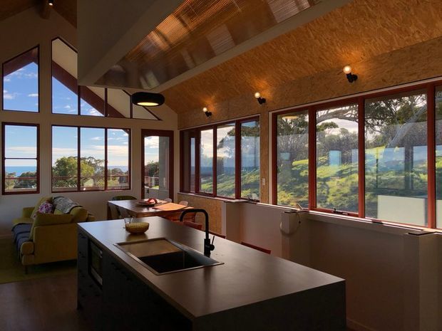 A warm, contemporary open-plan kitchen and living area with a large central island, integrated sink and dark cabinetry. Extensive timber-framed glazing wraps around the room, providing views toward a coastal or rural landscape. The room has a distinctive angled ceiling lined with textured timber panels, exposed roof structure, and warm recessed and pendant lighting.