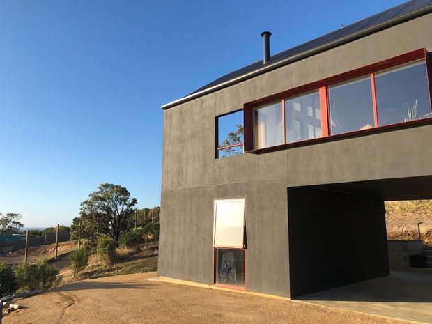 Exterior view of a contemporary two-storey home with a dark textured rendered facade, low-pitched roof, and strong horizontal glazing framed in reddish timber or metal. A deep recessed ground-floor opening creates a covered entry or carport-like void. The house sits on a dry, sloping landscaped site with distant coastal or rural views.