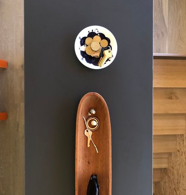Top-down view of a dark kitchen island or benchtop with a white bowl of fruit and keys, alongside a long timber serving tray holding a coffee cup and spoon. Warm timber cabinetry and flooring are visible at the sides, with small orange drawer pulls adding a bright accent.