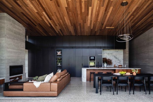 Open-plan contemporary living and dining area with a dark built-in kitchen, large marble island, brown leather sectional sofa, black dining furniture, polished concrete walls, terrazzo-like flooring, and a richly timber-lined ceiling. A sculptural pendant light hangs above the dining table, creating a refined, warm, high-contrast interior.