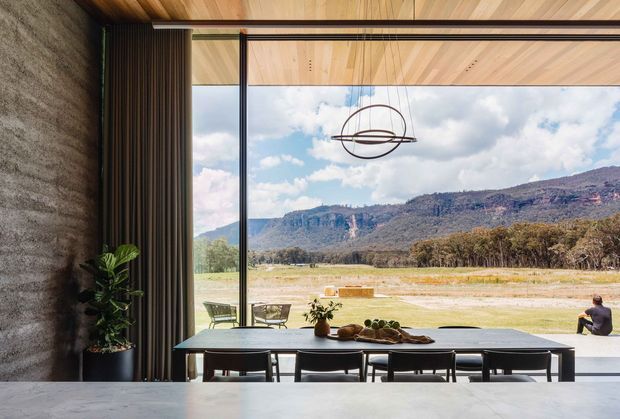 Contemporary dining area opening through expansive floor-to-ceiling glazing to a broad rural landscape and mountain escarpment. The room features a long dark dining table with upholstered chairs, a sculptural circular pendant, a textured concrete wall, timber ceiling and cabinetry or wall screening, and a large open glass wall creating a strong indoor-outdoor connection.