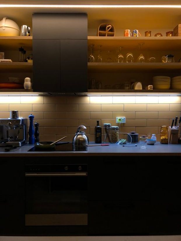 A compact, moody contemporary kitchen with dark matte cabinetry, a grey benchtop, warm brown subway-tile splashback, open timber-toned shelving, under-cabinet lighting, and integrated appliances. The shelves display glassware and kitchenware, while the benchtop includes a kettle, coffee machine, cookware, and utensils.