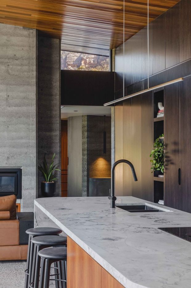A sophisticated open-plan kitchen viewed from the island, featuring a large veined stone island benchtop with an integrated sink and matte black gooseneck tap, warm timber cabinetry, dark vertical-panel cabinetry, and dark stone or concrete wall surfaces. Bar stools line the island, with a leather sofa and fireplace visible at left. The space has a high timber-lined ceiling, expansive upper glazing, and a dramatic mix of natural timber, concrete, and dark finishes.