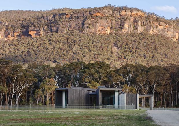 A low, modern single-storey house set in an open grassy clearing at the edge of eucalyptus woodland, with a dramatic layered sandstone cliff rising behind it. The dwelling has dark vertical cladding, extensive floor-to-ceiling glazing, crisp rectangular volumes, and a covered entrance or carport-like structure. The setting is rural and bushland-oriented, with a restrained contemporary architectural character.