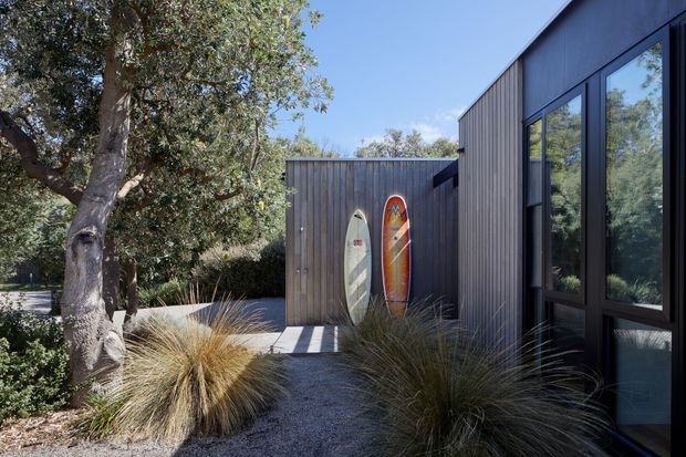 Exterior view of a contemporary dark-clad building set among established trees and ornamental grasses. A gravel walkway leads to a timber-clad section with two surfboards displayed against the wall, while large dark-framed windows reflect the surrounding vegetation. The scene has a relaxed, nature-connected residential character.