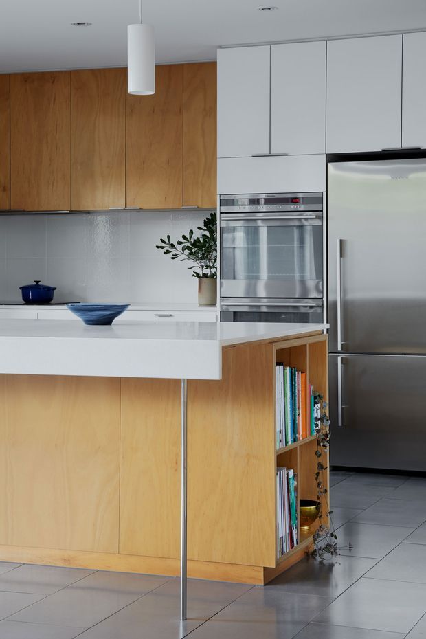 A contemporary kitchen with warm timber cabinetry, white upper cabinets, a large white island benchtop, integrated stainless-steel double ovens, and a stainless-steel refrigerator. The island includes open timber shelving for cookbooks and decor, with pale grey tiled flooring, a white cylindrical pendant light, and a light grey backsplash. A small plant and blue ceramic vessels add restrained accents.