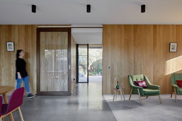 A refined mid-century-inspired living room or entry lounge with full-height vertical timber wall panelling, a smooth white ceiling with black recessed cylindrical spotlights, and large grey tiled flooring. A dark-framed glazed sliding door and central hallway open toward an outdoor area. Green upholstered lounge chairs with timber legs and burgundy cushions sit on a grey rug, while framed artwork and a small side table add restrained accents. Strong natural light enters from the right, creating a warm, calm interior with a clear view through to the exterior.