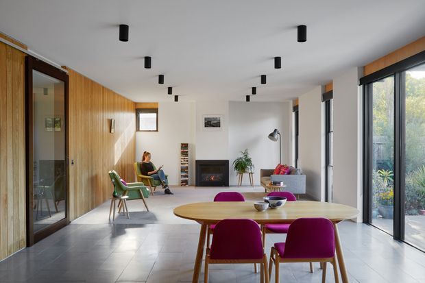 A bright, open-plan contemporary interior combining dining and living areas. The room features a long timber-lined wall with dark-framed glazed doors, pale tiled flooring, white walls and ceiling, a compact fireplace, large black-framed windows, and a timber dining table with vivid magenta upholstered chairs. A seating area, reading nook, plants, and contemporary lighting complete the space.